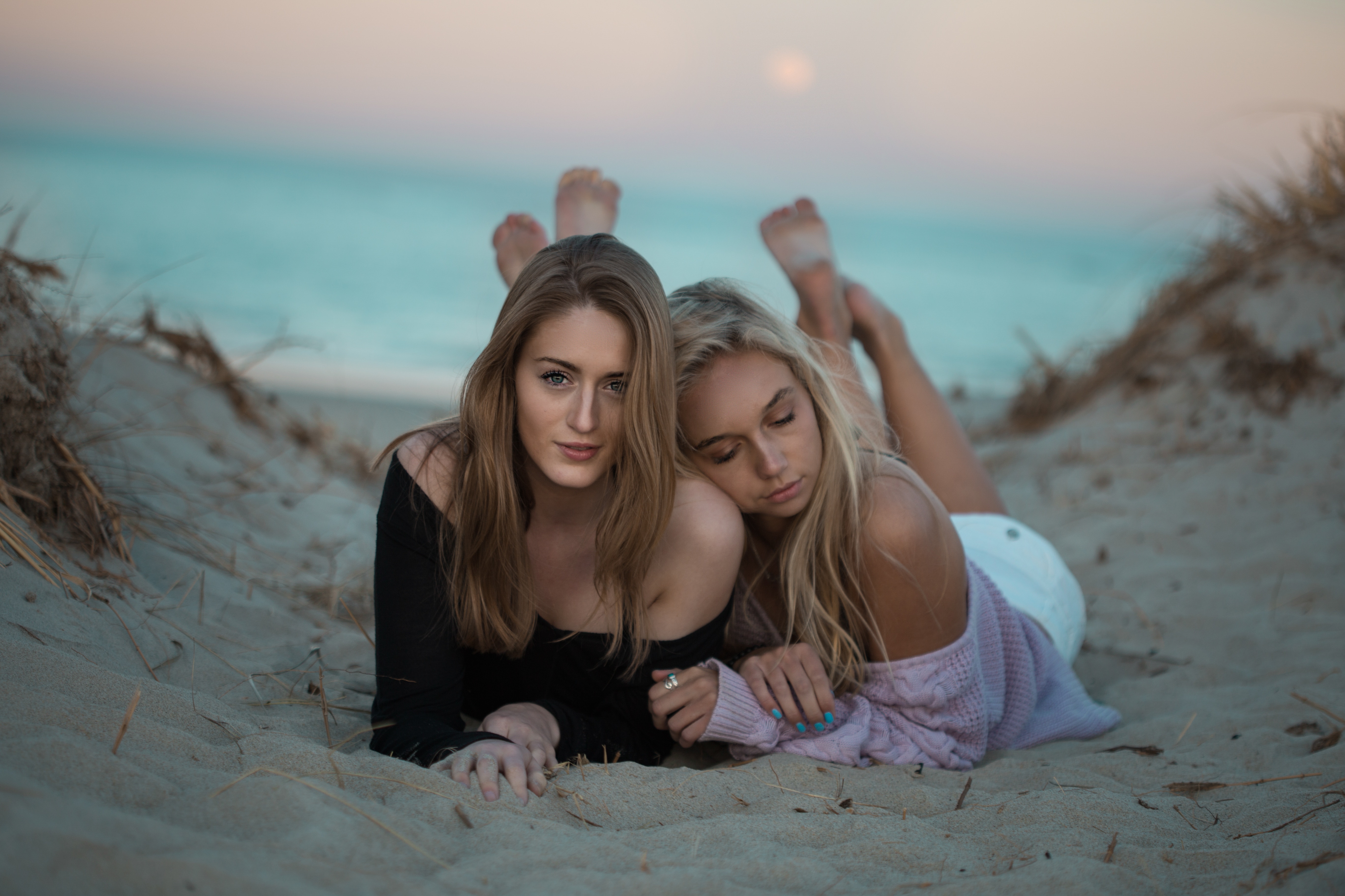 People 5472x3648 women two women long hair brunette blonde beach women outdoors sand lying on front nature bare shoulders model Kristopher Roller sea sunset closed eyes feet in the air caressing legs up barefoot depth of field