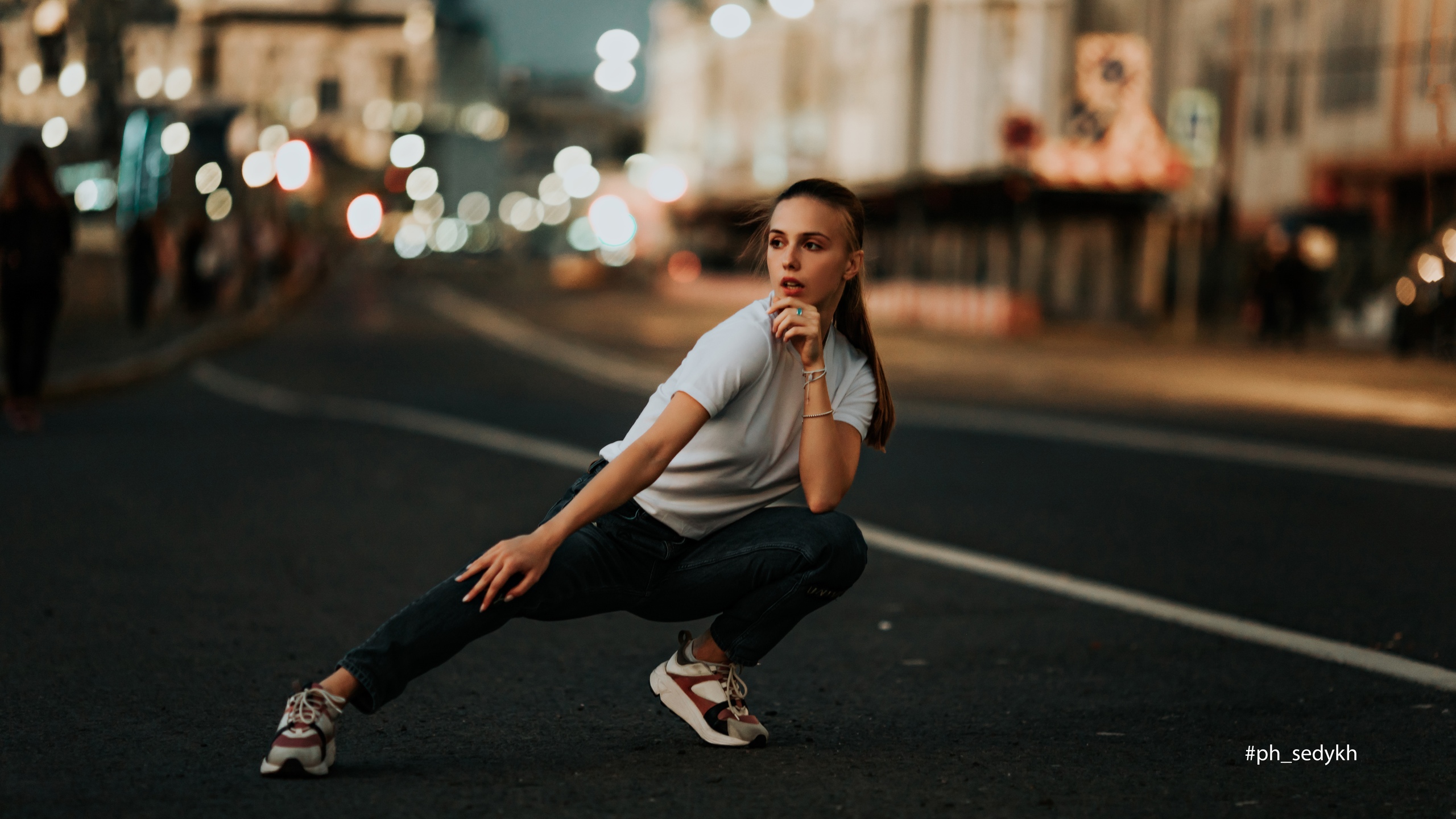 People 2560x1440 women brunette ponytail T-shirt jeans sneakers looking away touching face squatting street urban city lights outdoors women outdoors Dmitry Sedykh watermarked
