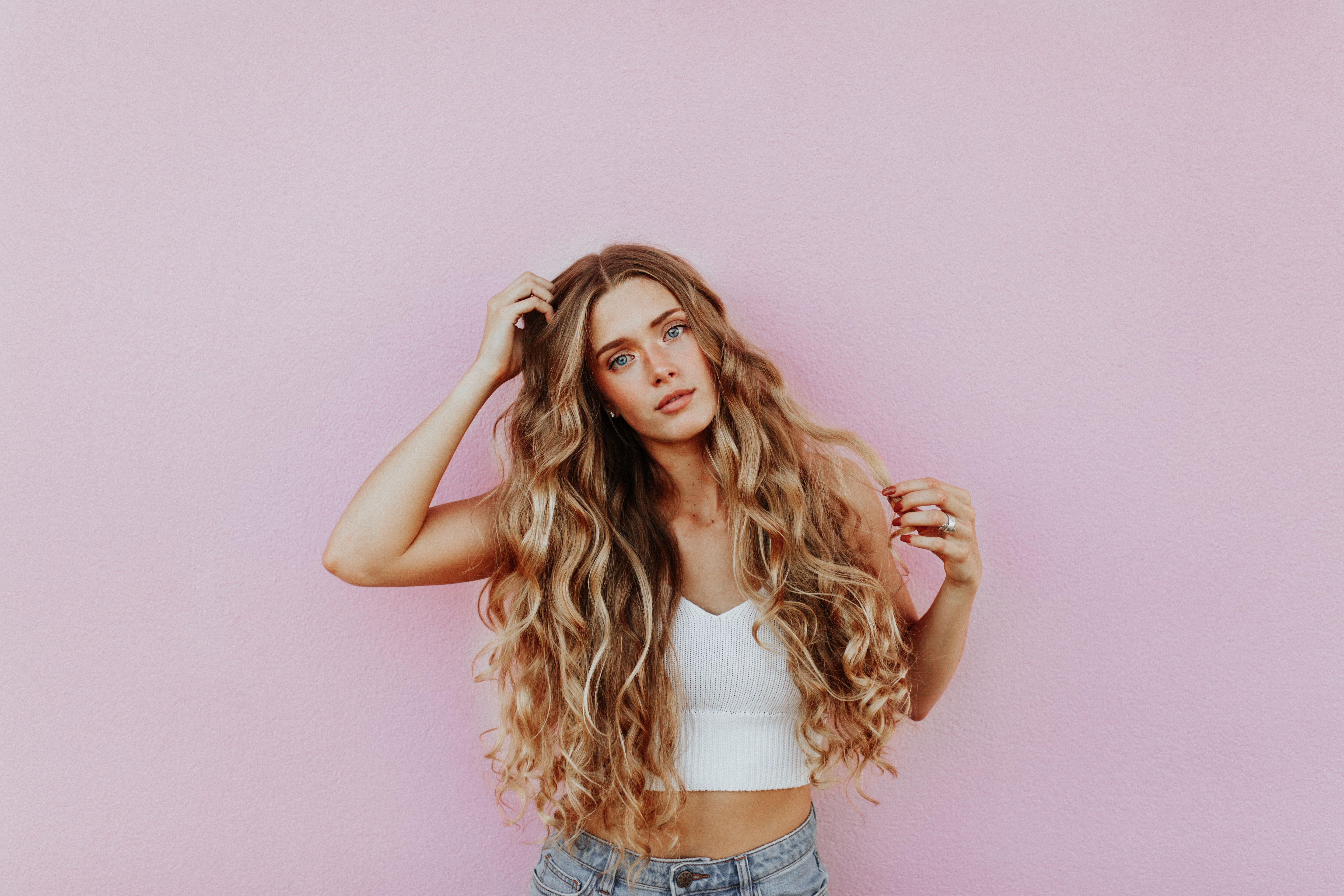 People 5760x3840 photography women model women indoors face jean shorts belly long hair simple background pink background curly hair hands in hair tank top blue eyes looking at viewer