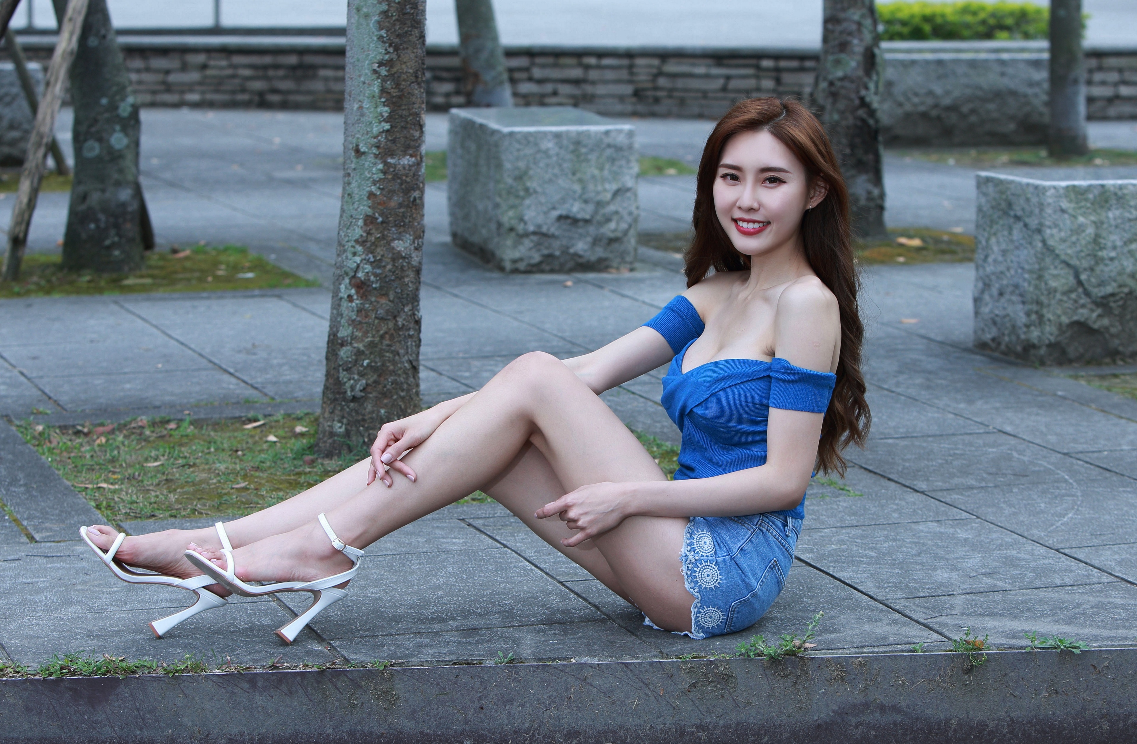 People 3840x2514 Asian model women long hair dark hair sitting barefoot sandal jean shorts blue tops bare shoulders trees stones grass bushes depth of field white heels short shorts smiling red lipstick looking at viewer outdoors dyed hair legs