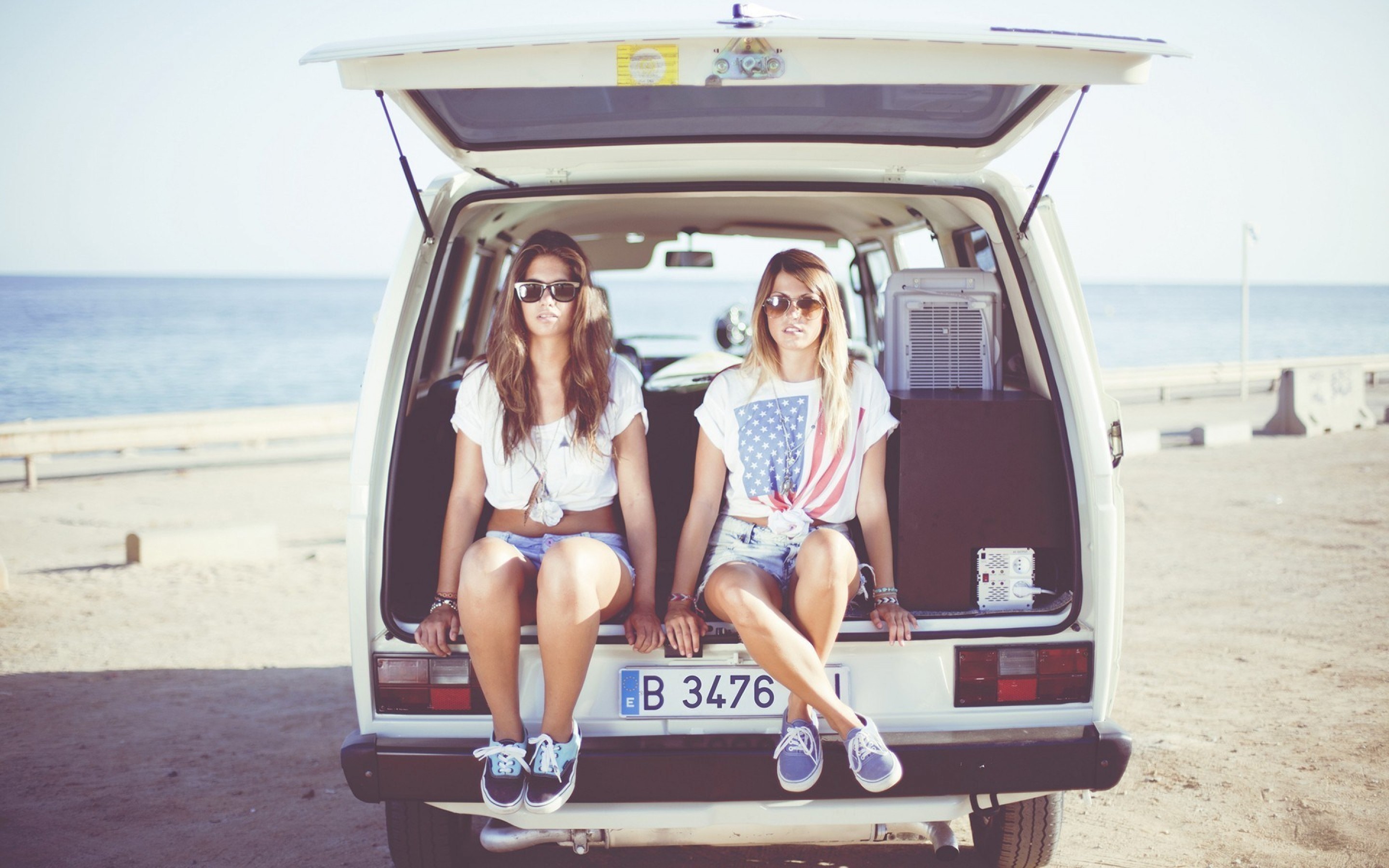 People 2880x1800 women model brunette blonde long hair straight hair face sunglasses T-shirt shorts jean shorts Volkswagen sitting beach van legs looking at viewer Spain Volkswagen Transporter frontal view sea no socks sneakers feet crossed tied top short sleeves bent legs