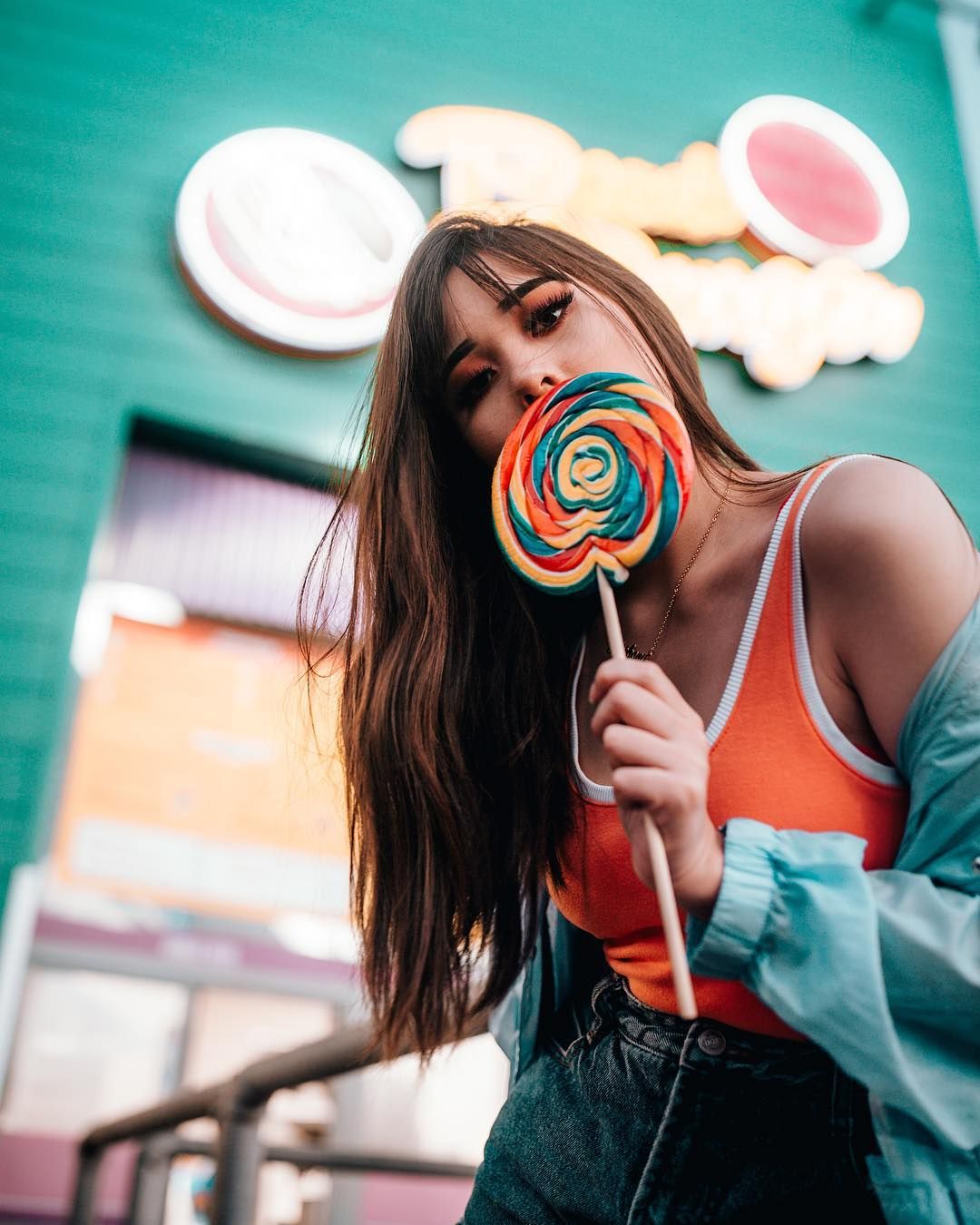 People 1080x1350 women model brunette long hair straight hair eyeshadow lollipop jacket women outdoors portrait display bokeh natural light looking at viewer tank top jeans orange clothing leaning hair in face arched back necklace colorful green food sweets makeup urban